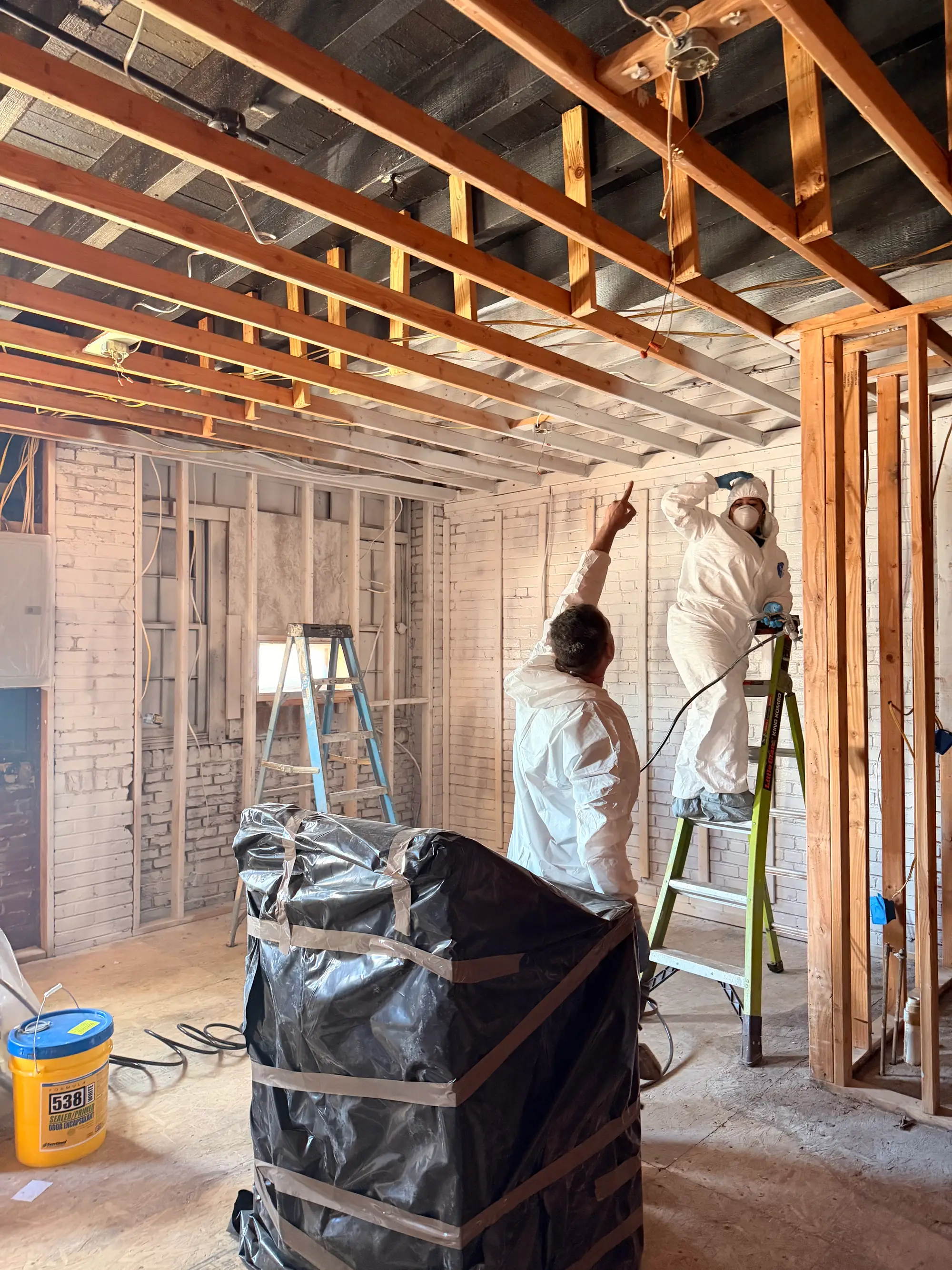 Two technicians in Tyvek suits at stripped wall framing with sealed disposal bags during mold remediation
