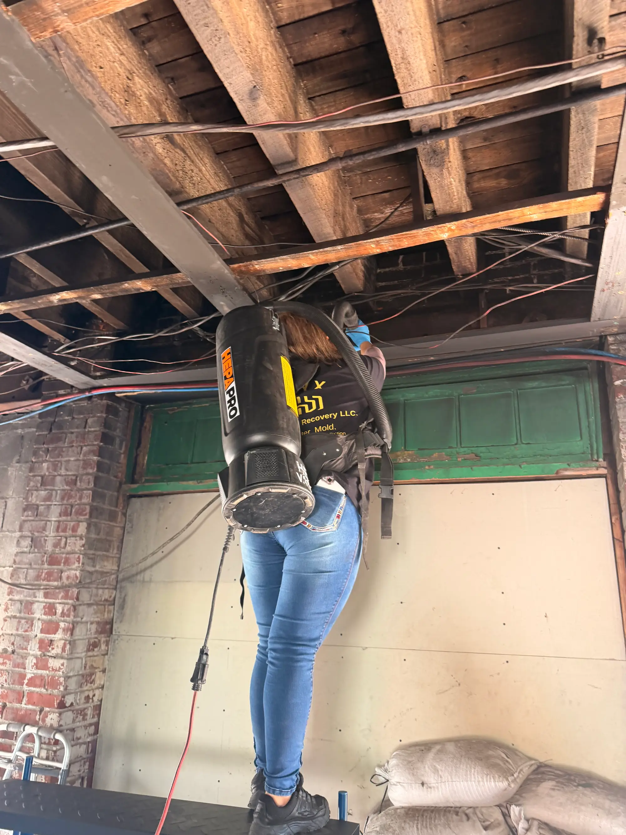 Technician using a HEPA backpack vacuum on exposed joists during mold removal